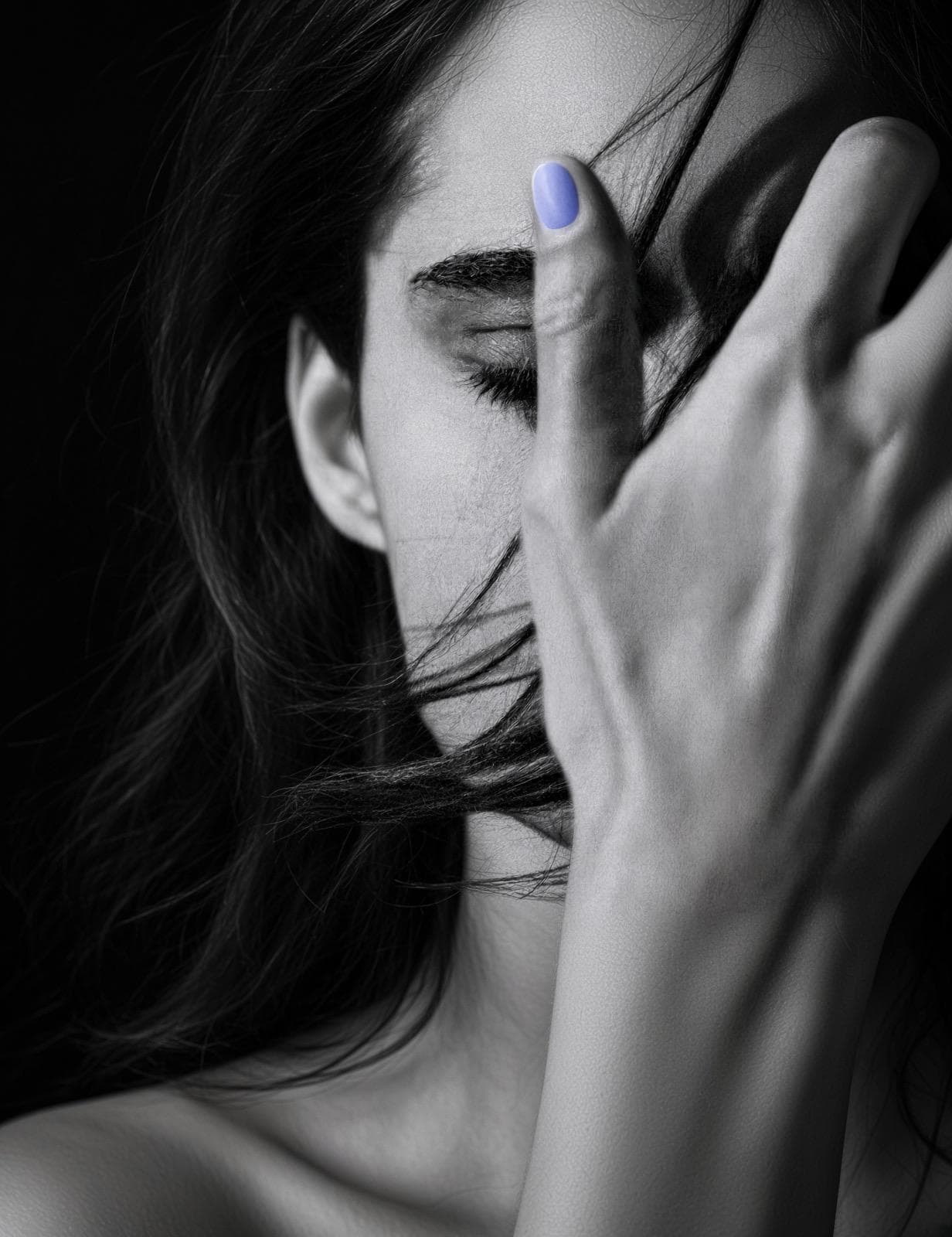 A woman's face behind her own hand, fingers spread across it, eyes closed beneath them, hair crossing the frame, in black and white with a single painted nail in soft periwinkle.