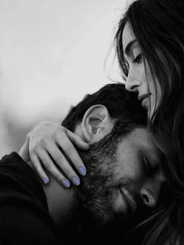 Two people close together on a pale ground, one resting against the other's shoulder with eyes closed, a hand holding them steady, in soft light, the nails carrying a single touch of soft periwinkle.