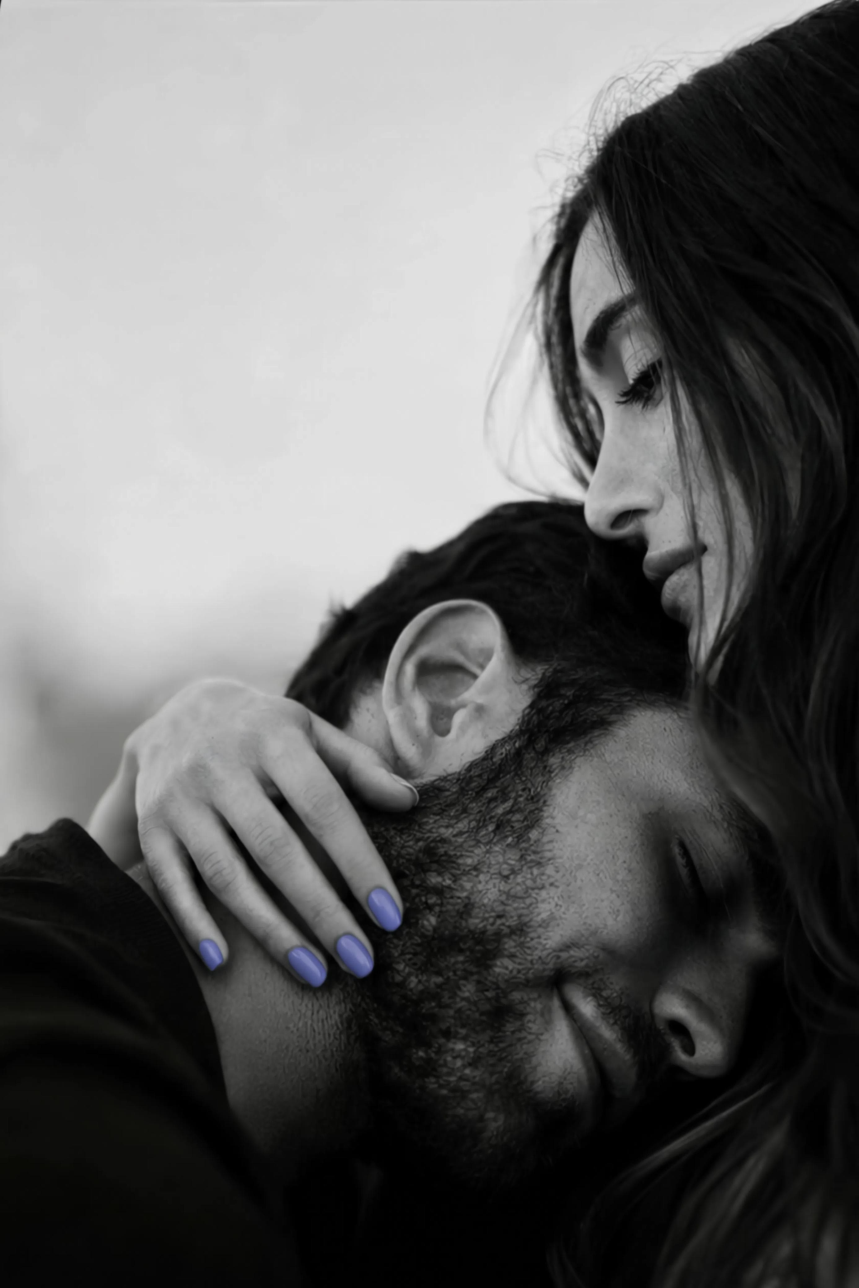 Two people in a close embrace, in black and white, one head resting against the other and a hand holding it there, the nails carrying a single touch of soft periwinkle.