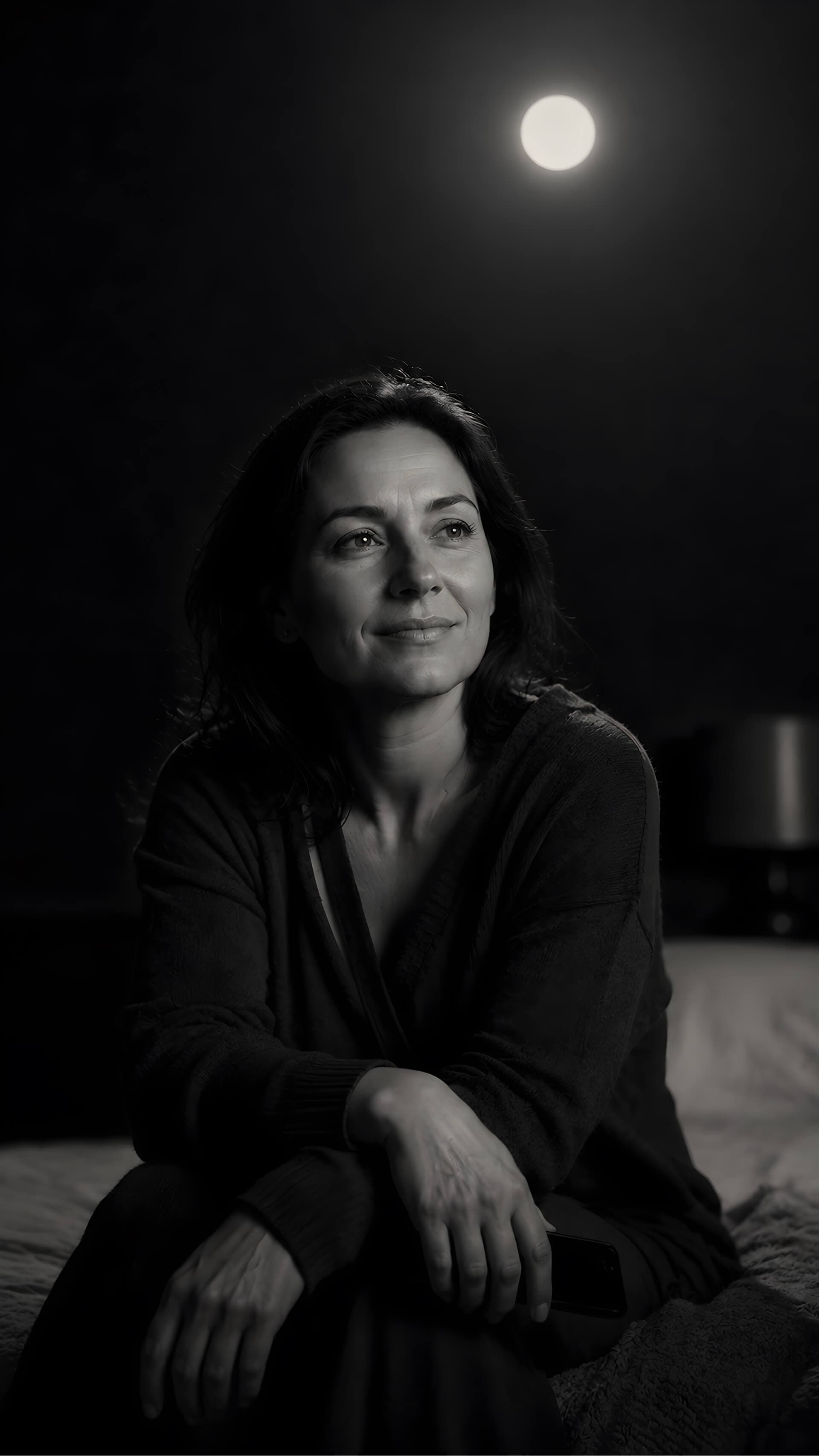 A figure sitting on a bed in a dark room, lit by a small round source of warm light high on the wall behind, the rest of the room falling away into black.