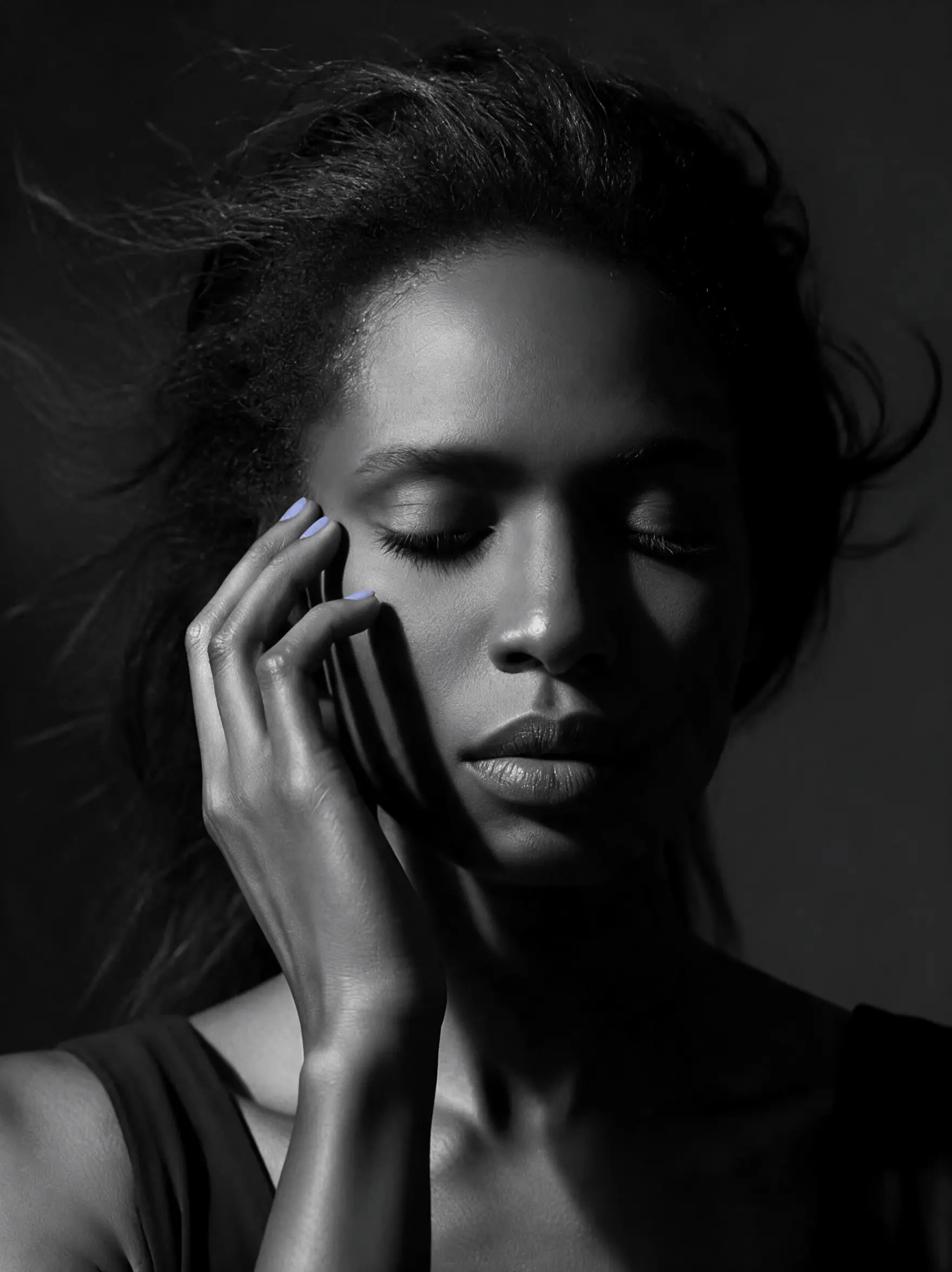 A woman's head and shoulders against a deep charcoal ground, head tilted down and eyes closed, one hand resting at her temple, hair caught mid-movement, lit softly from the upper left.