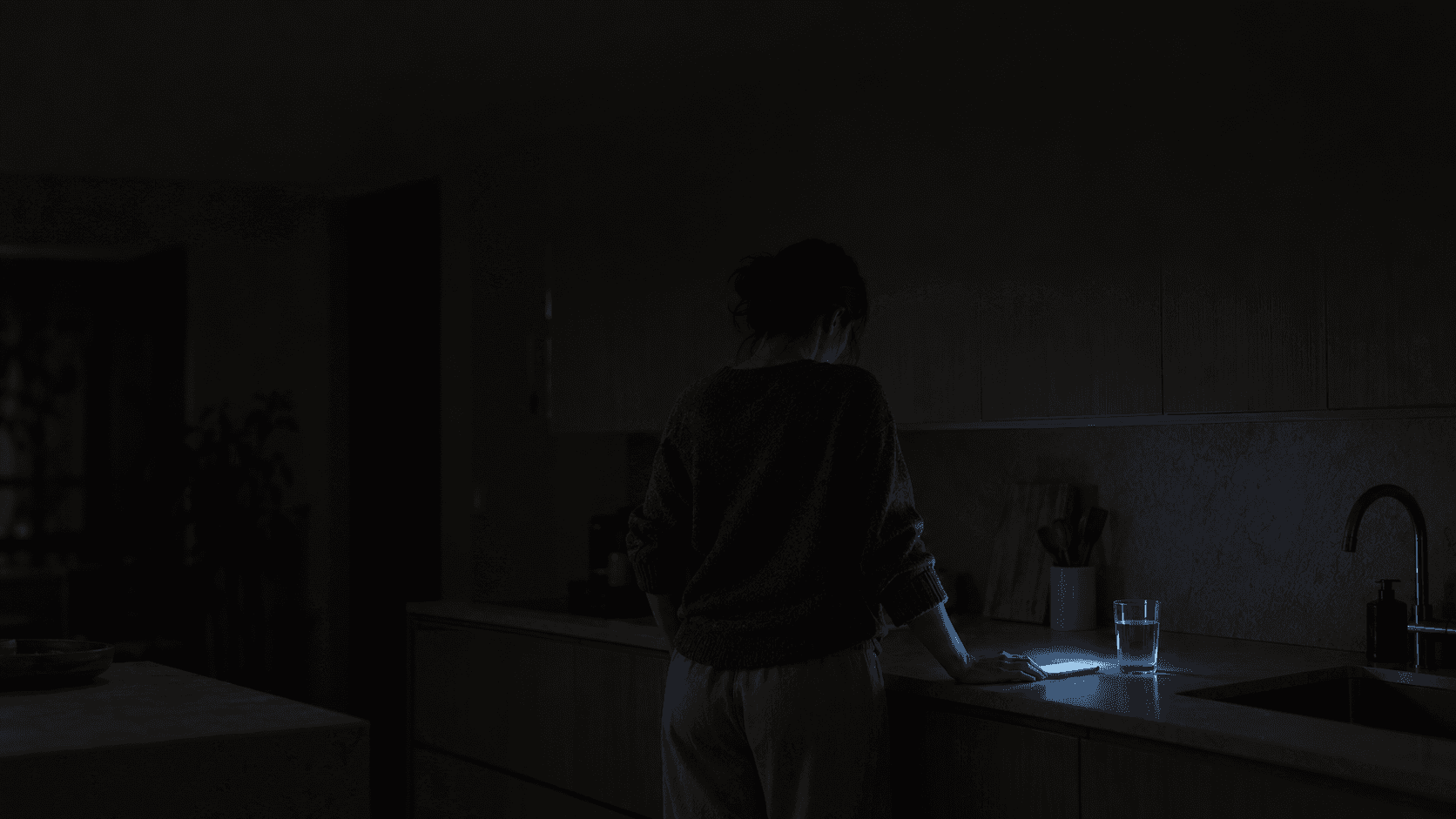 Woman awake alone in a dark kitchen with a phone glowing on the counter, illustrating the checking pattern described in relationship OCD.