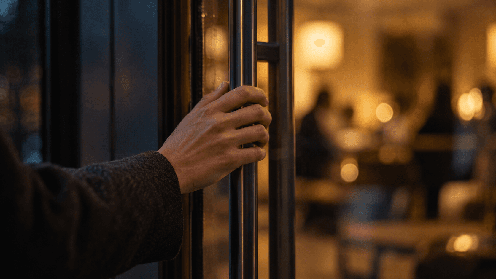 A hand resting on a glass door handle with warm light and blurred figures in the room beyond