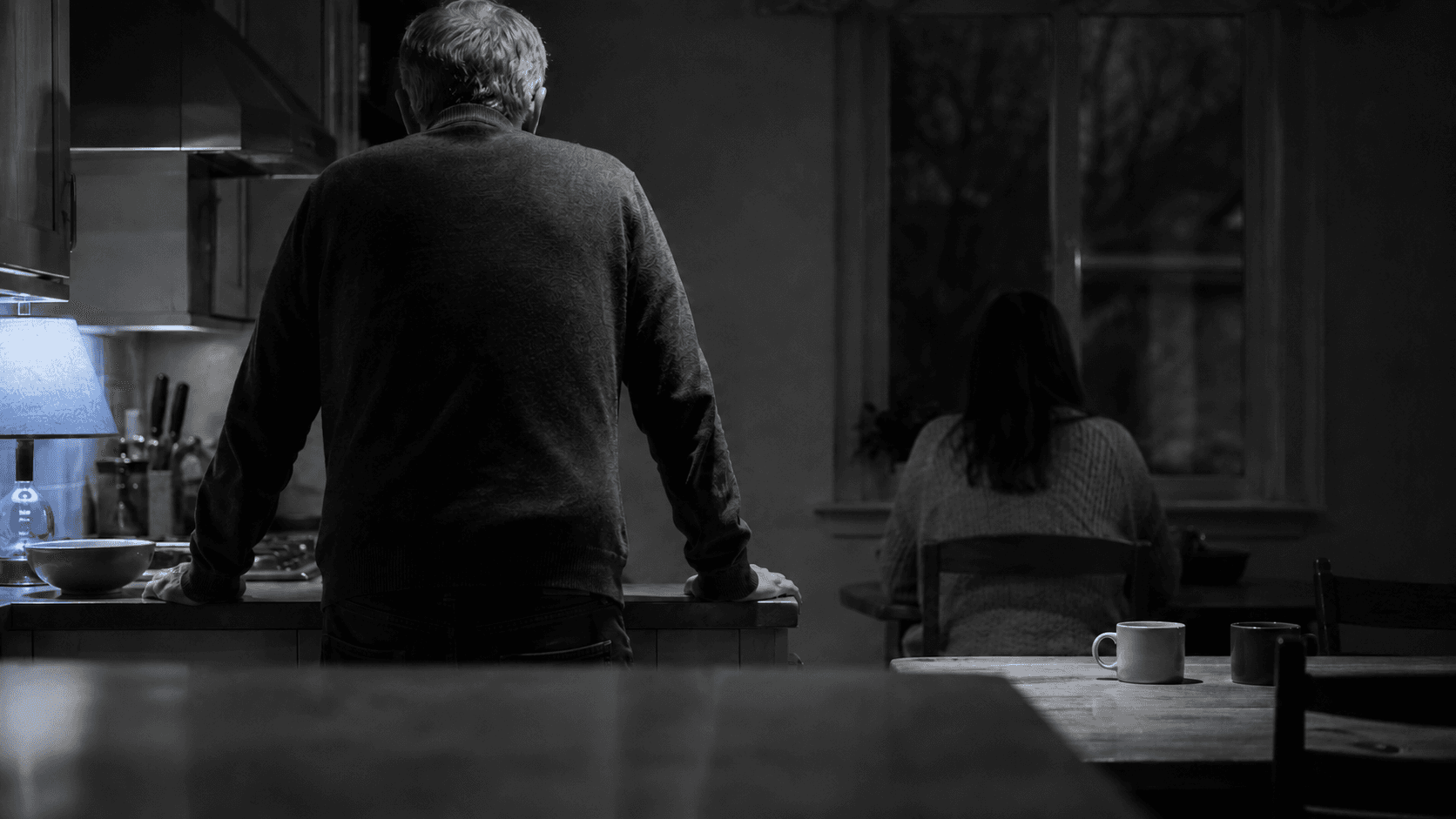 Older man and younger woman apart in a dim kitchen at night, illustrating the distance in age gap relationships