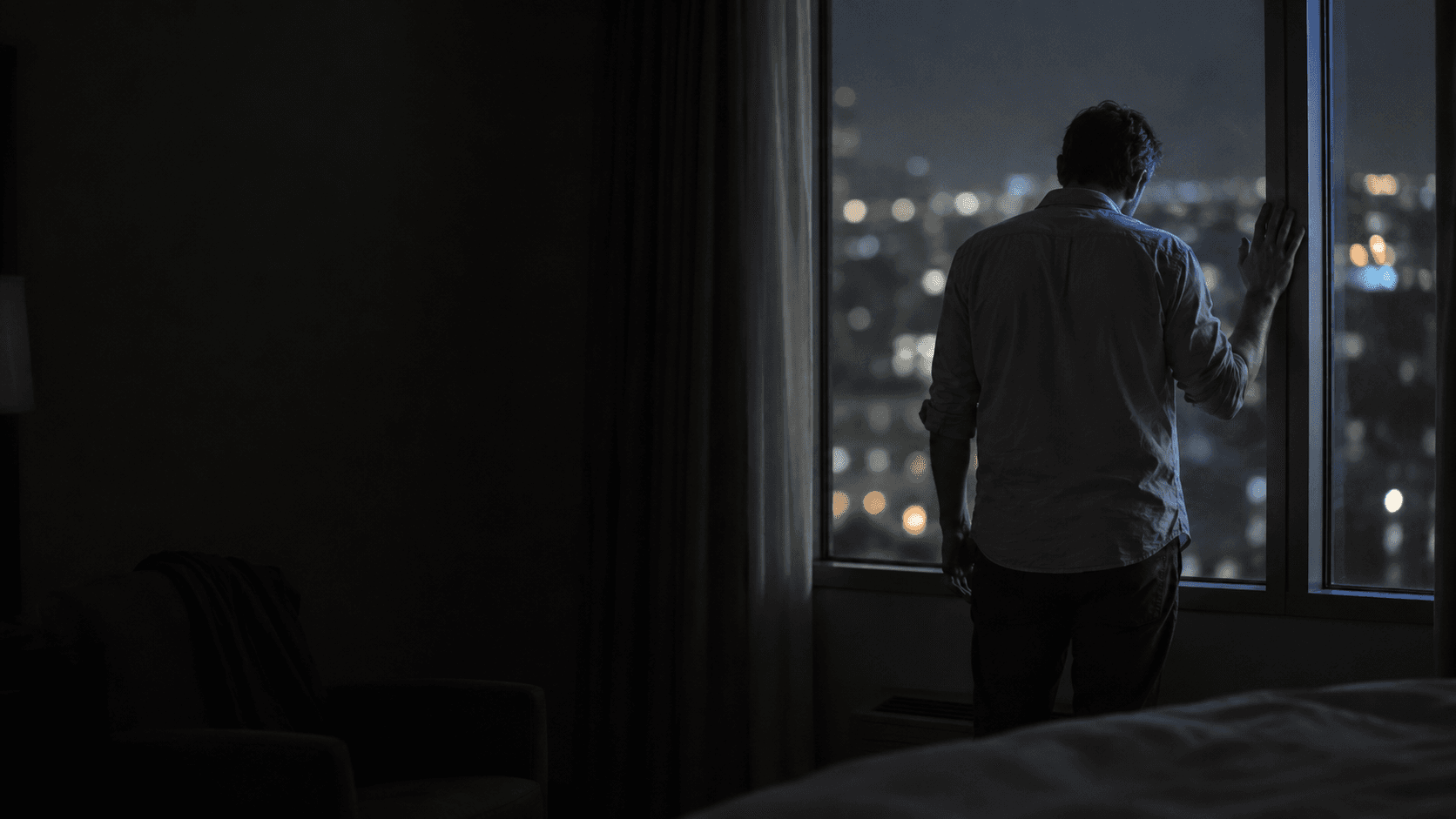 Man standing alone at a hotel window at night with the room dark behind him, illustrating male infidelity patterns