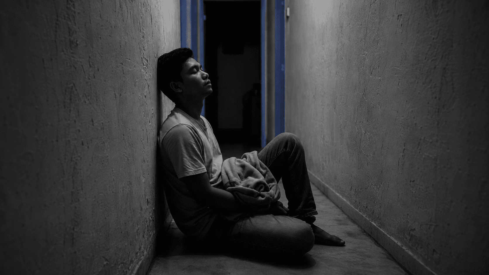 A man sitting alone in a dark hallway at night, reflecting how to regain trust after being cheated on, with pale blue light in the doorway behind him.