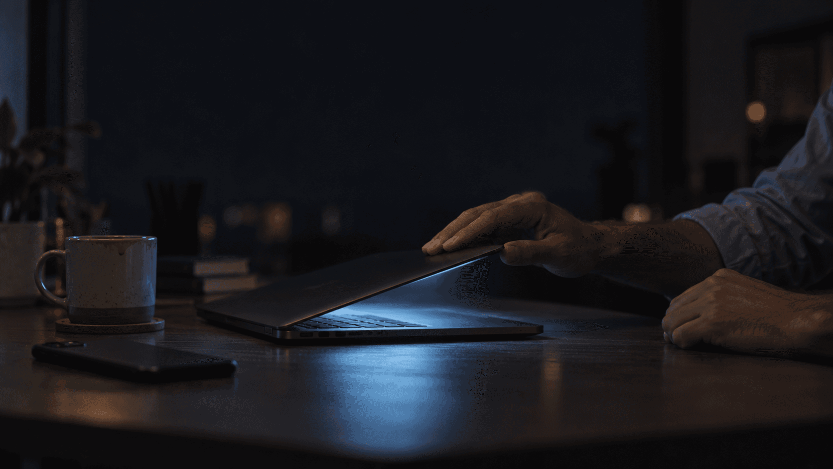 A man's hands closing a laptop in a dark room late at night, phone face down beside it