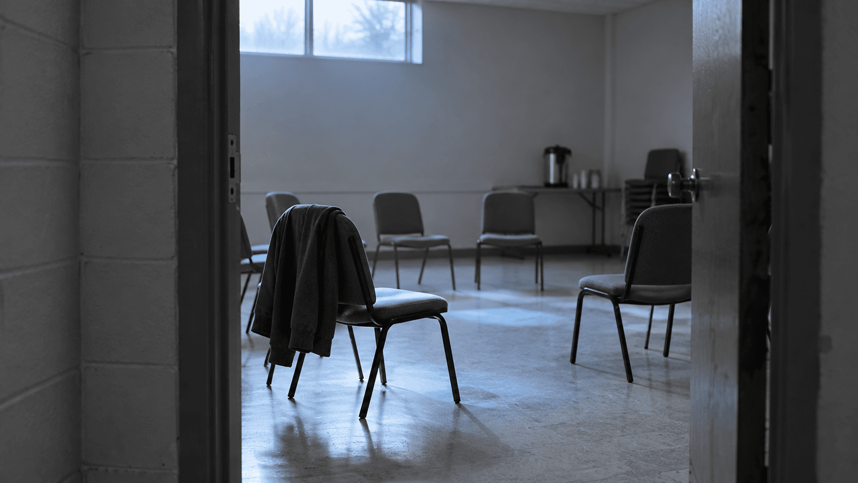 An open door into a community meeting room with a circle of empty chairs, a coat over the nearest one and a coffee urn on a table behind: widow and widower support groups near me