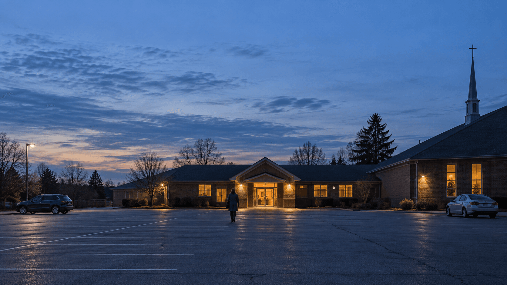 Person walking across a dusk parking lot toward a lit church hall for a divorce support meeting