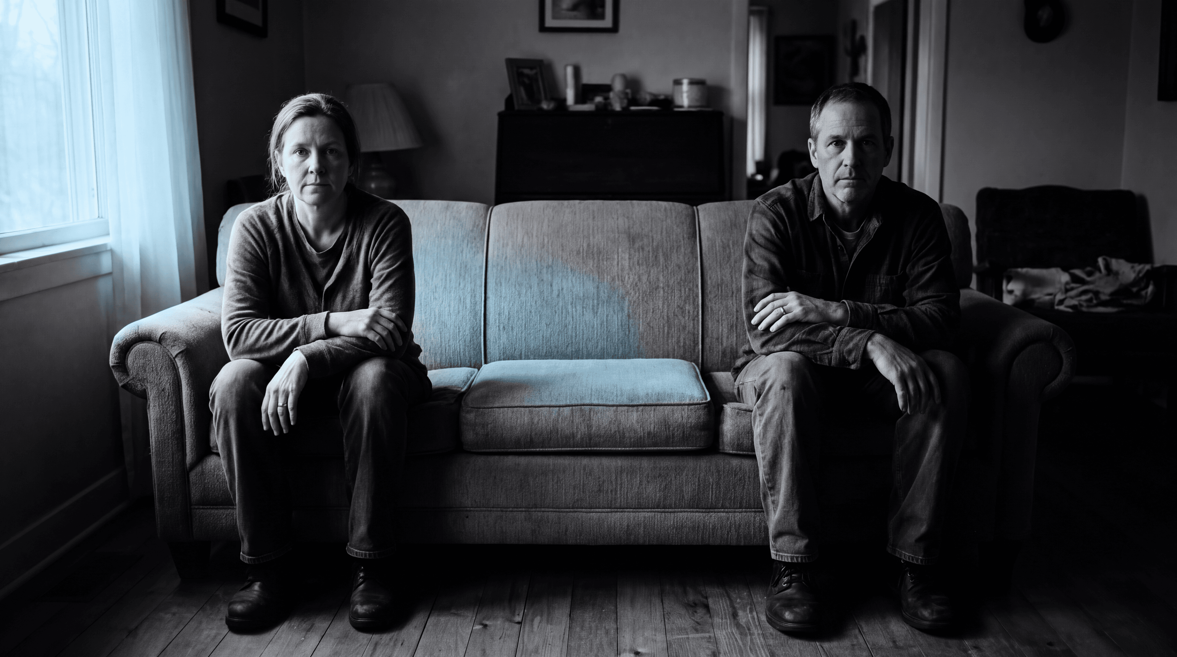 A married couple sits at opposite ends of a couch with an empty cushion between them, evoking the quiet distance of a sexless marriage.